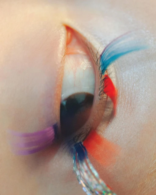Extreme close-up of a human eye with creative makeup in vibrant blue, red, and purple tones. Eyelashes are decorated with shimmering strands.