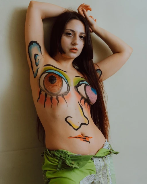A young woman with dark hair and whimsical body paint poses in front of a wall, looking at the camera with her arms up.