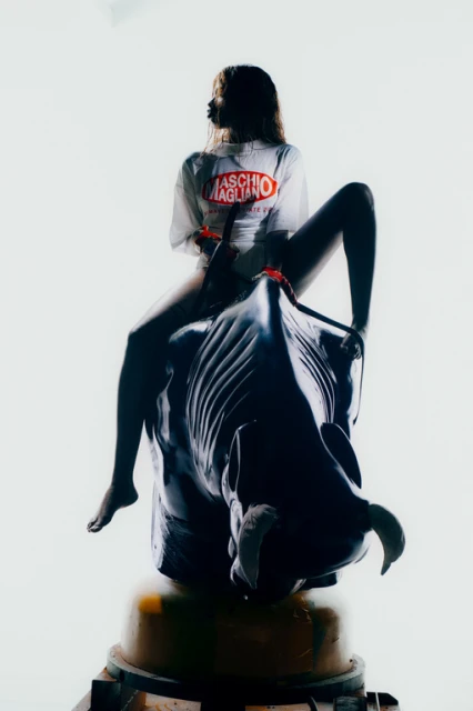 A person sits atop an artificial bull in a white top with Maschio Magliano text, creating a playful, fashion-forward image.