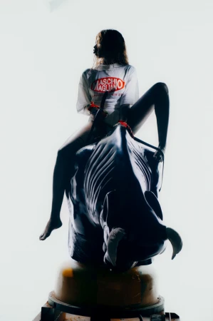 A person sits atop an artificial bull in a white top with Maschio Magliano text, creating a playful, fashion-forward image.