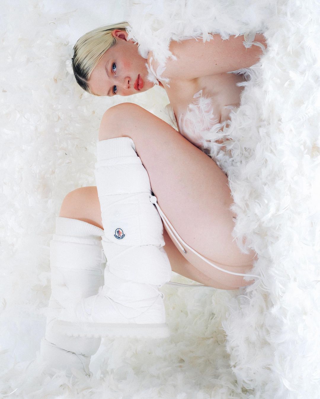 Woman poses lying among white feathers wearing white leg warmers, creating a surreal and fashionable fine art portrait.