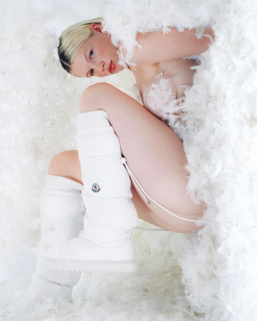Woman poses lying among white feathers wearing white leg warmers, creating a surreal and fashionable fine art portrait.