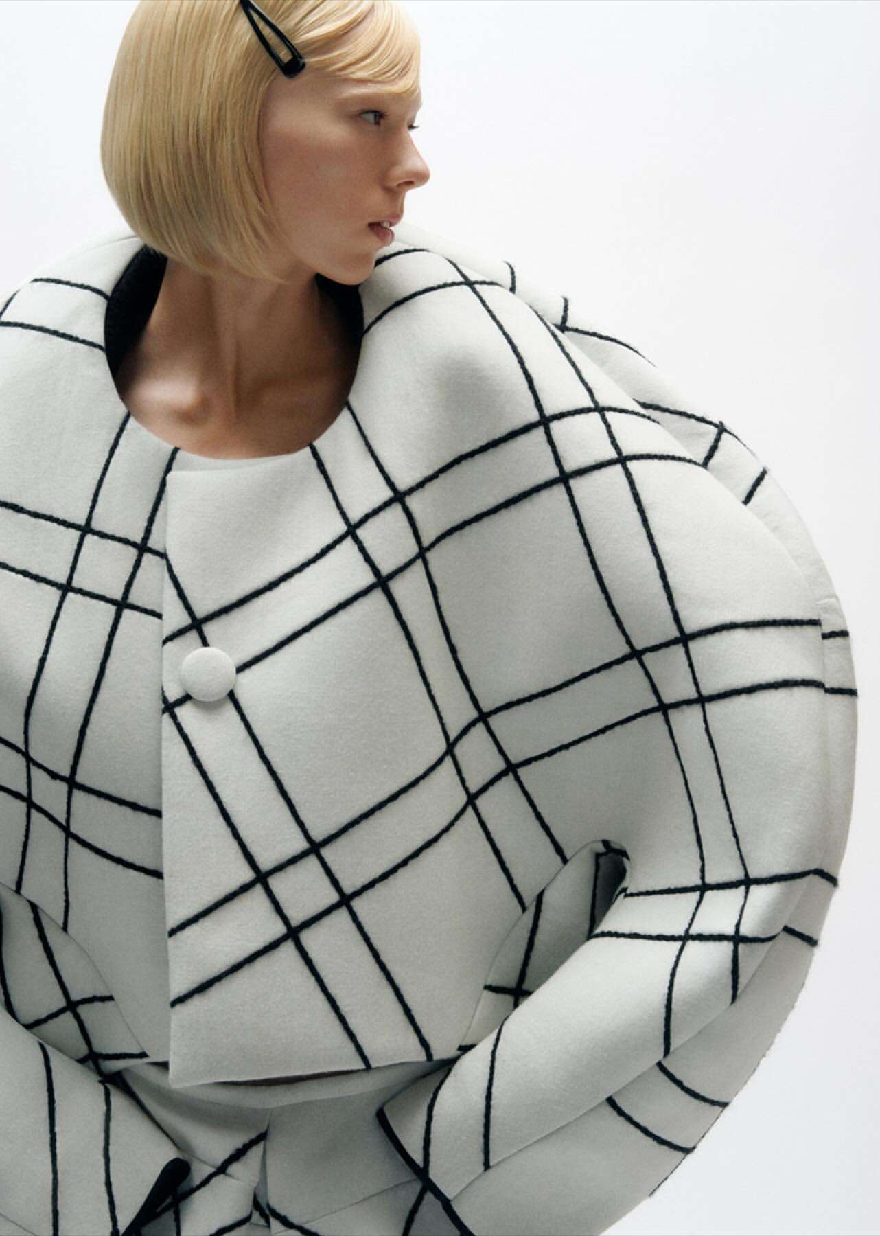 Fashion shot of a fair-skinned adult female with blonde hair wearing a white and black grid-patterned jacket and skirt against a white backdrop.