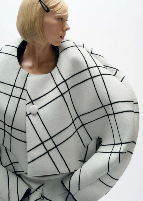 Fashion shot of a fair-skinned adult female with blonde hair wearing a white and black grid-patterned jacket and skirt against a white backdrop.