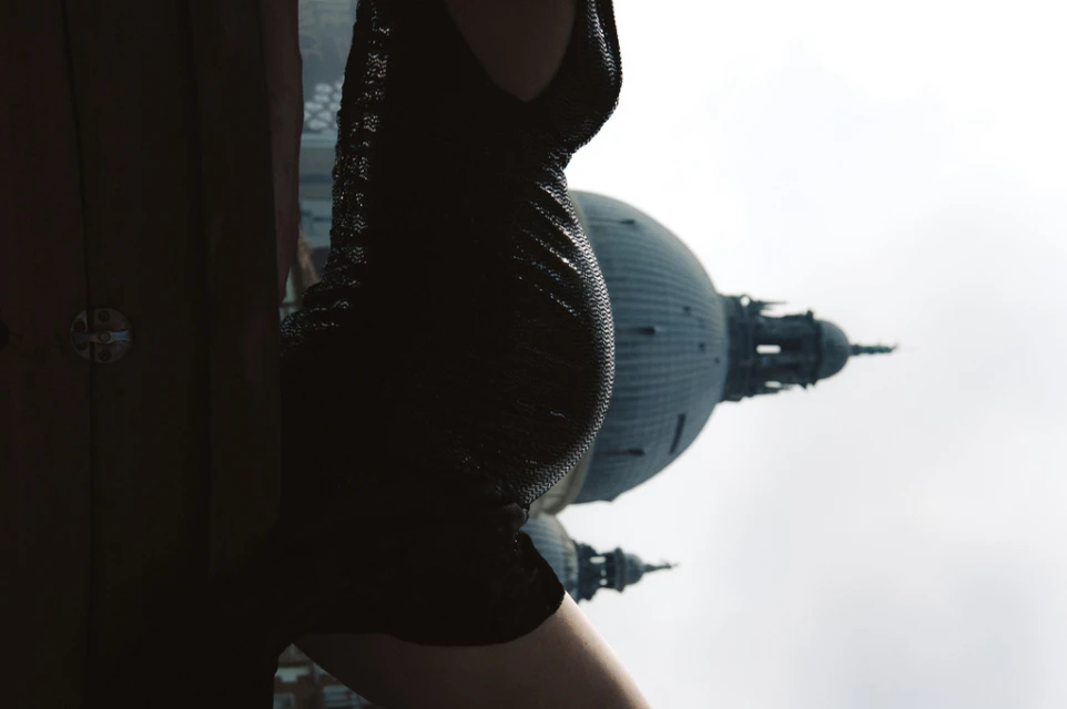 Woman poses in a black dress with architecture in the background during daytime with a hazy sky, creating a unique visual contrast.