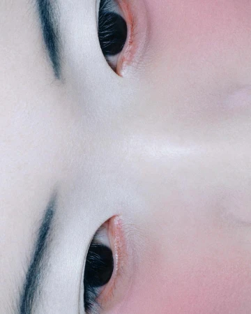 Close-up shot of a woman's face featuring dark eyes, black eyebrows, and a pale skin tone against a blurred pink background.