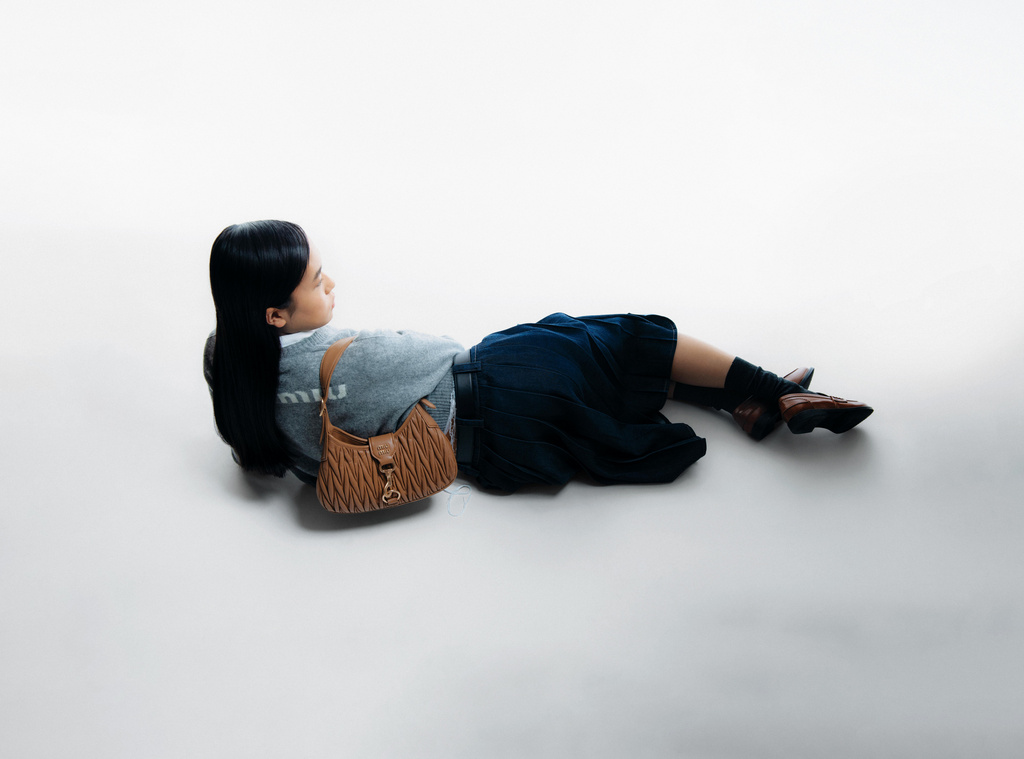 Fashion shot: young woman lying on a white background with a brown Miu Miu handbag, grey sweatshirt, and blue skirt in studio lighting.