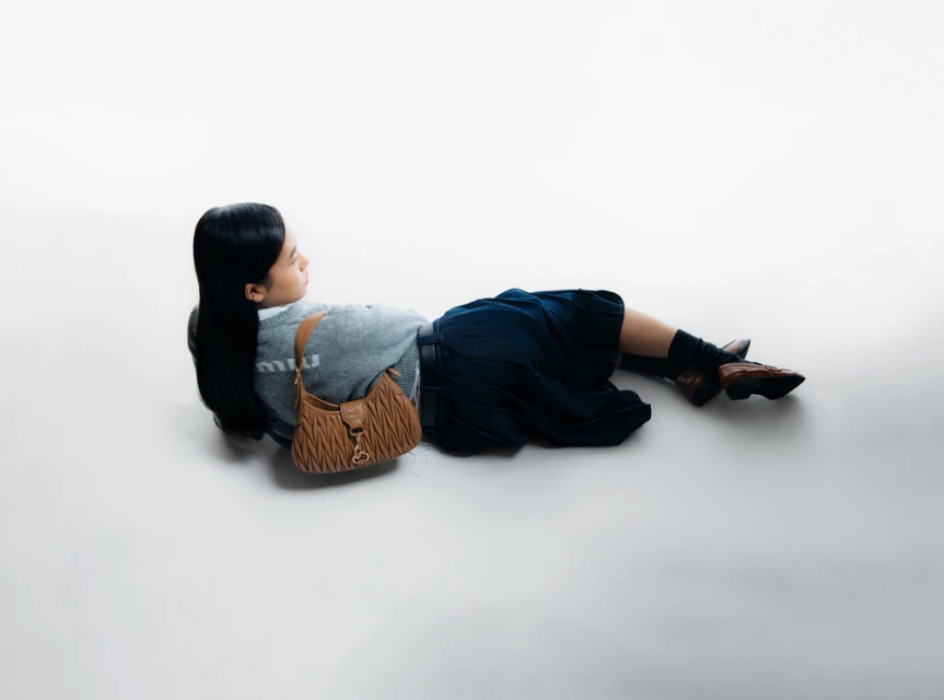Fashion shot: young woman lying on a white background with a brown Miu Miu handbag, grey sweatshirt, and blue skirt in studio lighting.
