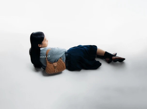 Fashion shot: young woman lying on a white background with a brown Miu Miu handbag, grey sweatshirt, and blue skirt in studio lighting.