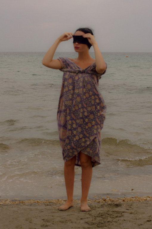 Adult female in a dress stands barefoot on a beach with arms raised and a blindfold covering her eyes near the ocean.