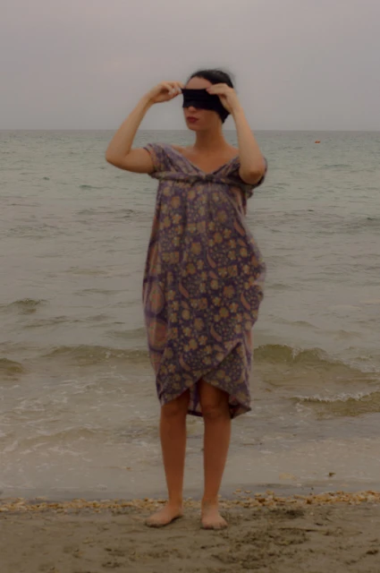Adult female in a dress stands barefoot on a beach with arms raised and a blindfold covering her eyes near the ocean.