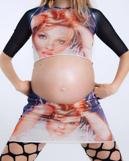 Pregnant woman poses in a mini-dress with a celebrity image printed on it, complemented by patterned stockings against a white backdrop.