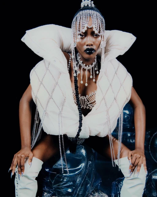 A striking fashion portrait of an African American woman in a cream-colored quilted vest, matching boots, and elaborate crystal jewelry on a black background.