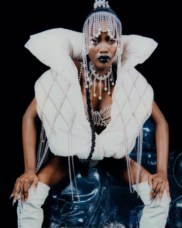 A striking fashion portrait of an African American woman in a cream-colored quilted vest, matching boots, and elaborate crystal jewelry on a black background.