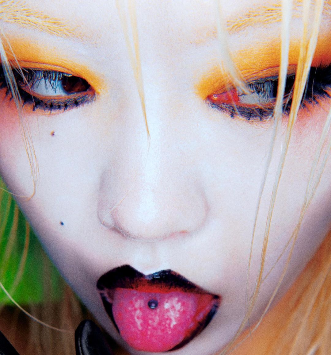 Close-up portrait of a fair-skinned person with blond hair and bright, colorful eyeshadow sticking their tongue out with a tongue piercing.