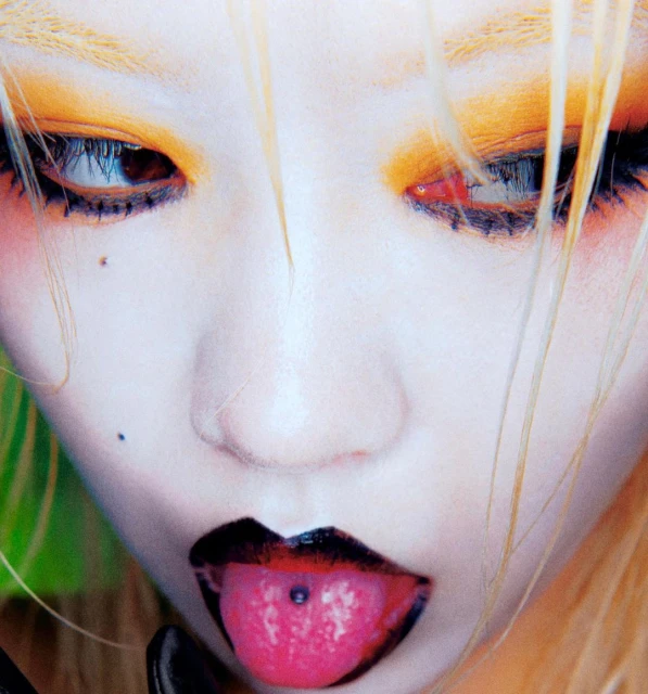 Close-up portrait of a fair-skinned person with blond hair and bright, colorful eyeshadow sticking their tongue out with a tongue piercing.