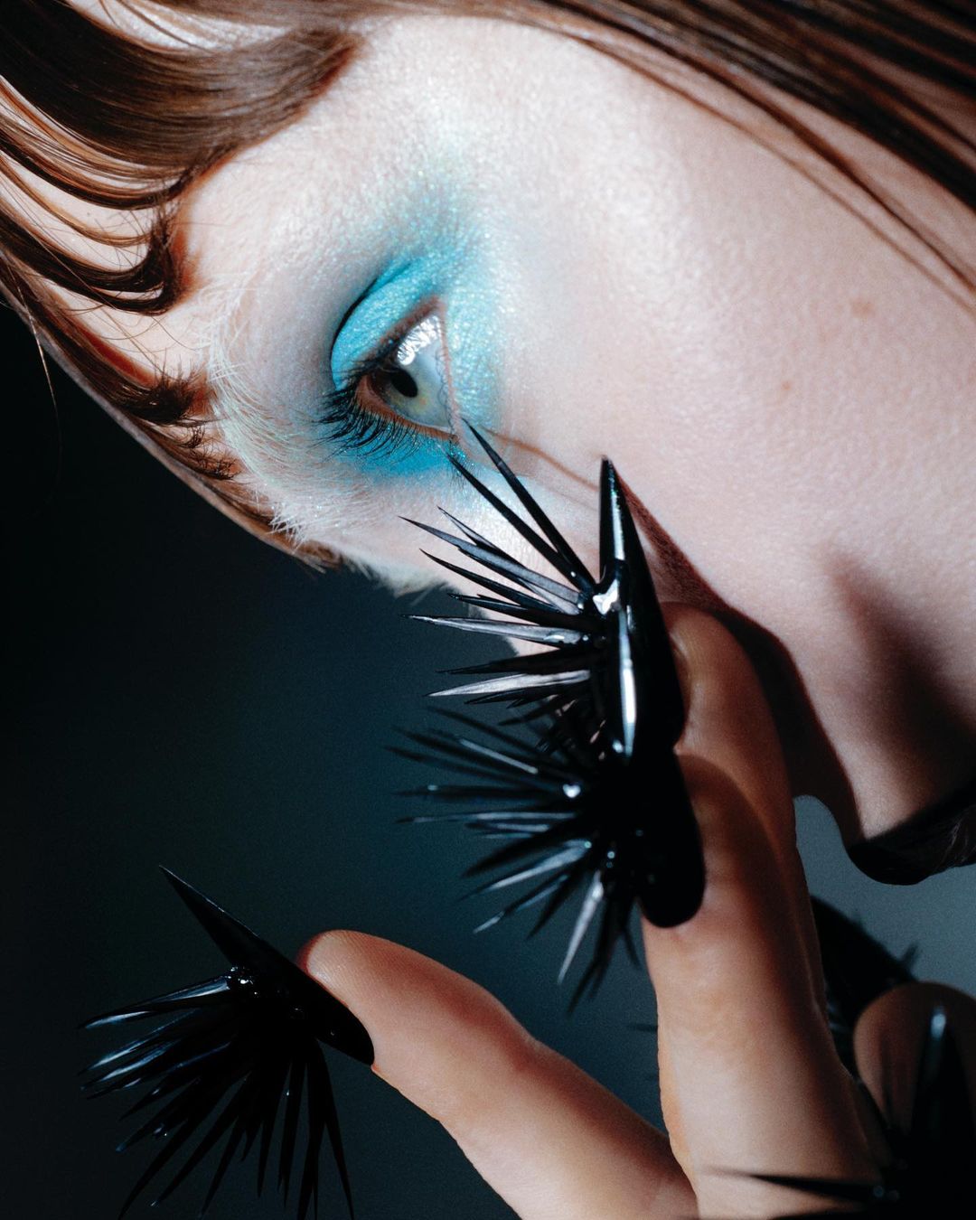 Close-up of a woman with blue eyeshadow and long, spiky black nail art. Studio lighting highlights the unique and dramatic makeup and nails.