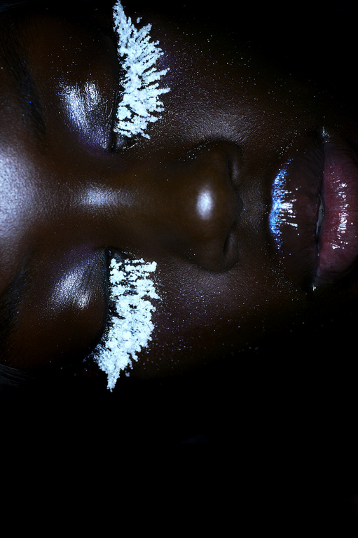 Close up portrait of a woman with dark skin, closed eyes, shiny skin, and white makeup on her eyelashes, and shimmering makeup on lips.