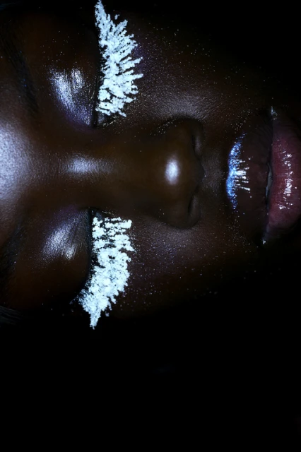 Close up portrait of a woman with dark skin, closed eyes, shiny skin, and white makeup on her eyelashes, and shimmering makeup on lips.