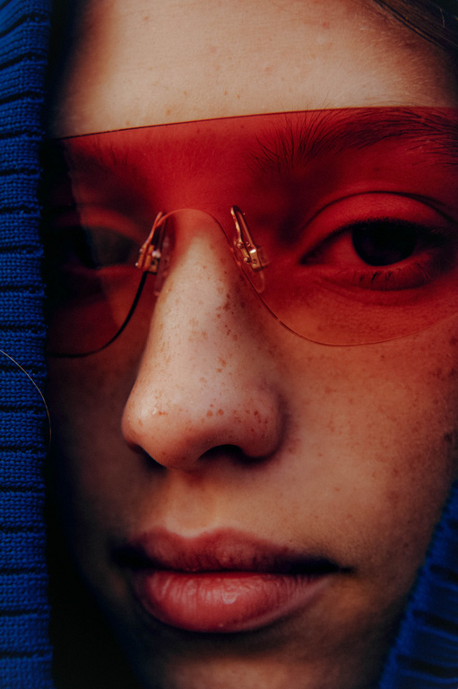 Close-up portrait of a person with freckles wearing translucent red sunglasses, blue hooded sweater, and neutral facial expression.