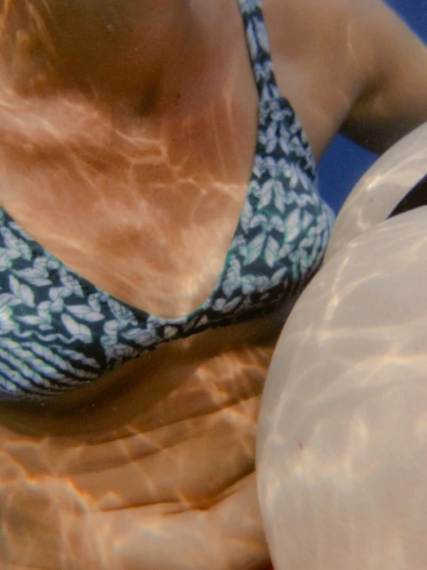 Close-up underwater shot of an adult woman wearing a bikini top, holding onto a white inflatable float. Water creates a rippling light effect.
