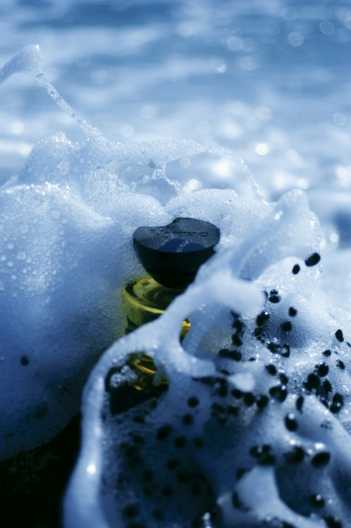 Close-up of a partially submerged glass bottle surrounded by seafoam on a sunny day, with the ocean visible in the background.