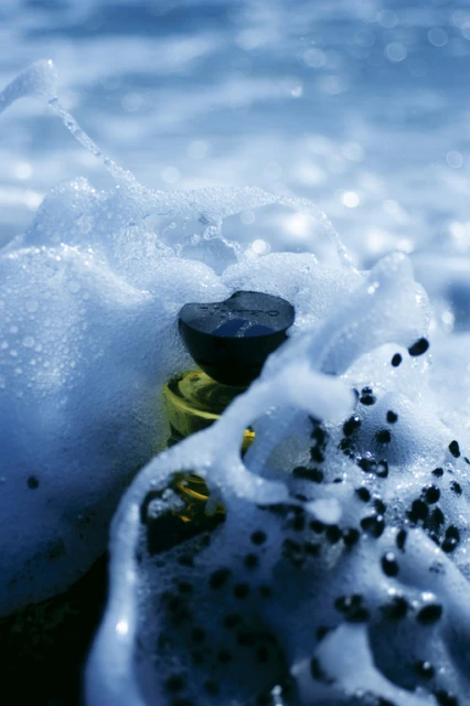 Close-up of a partially submerged glass bottle surrounded by seafoam on a sunny day, with the ocean visible in the background.