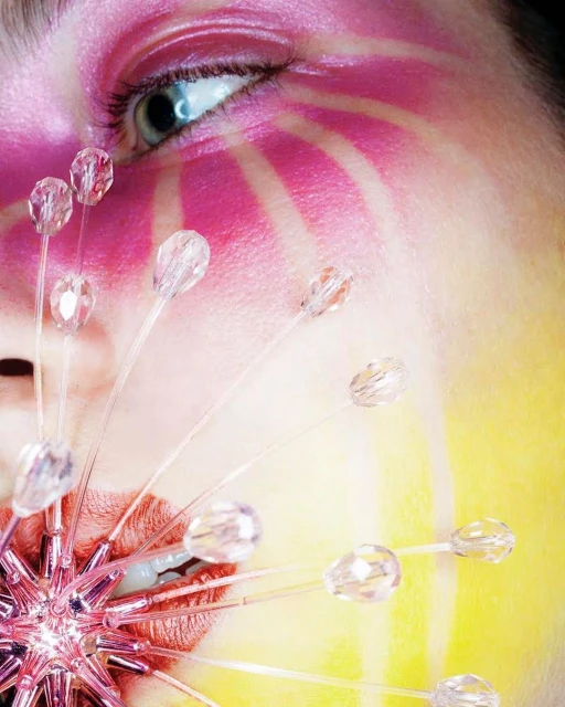 Close-up of a person with vivid pink eye makeup and radiating yellow facial paint. An accessory near the mouth features clear beads.