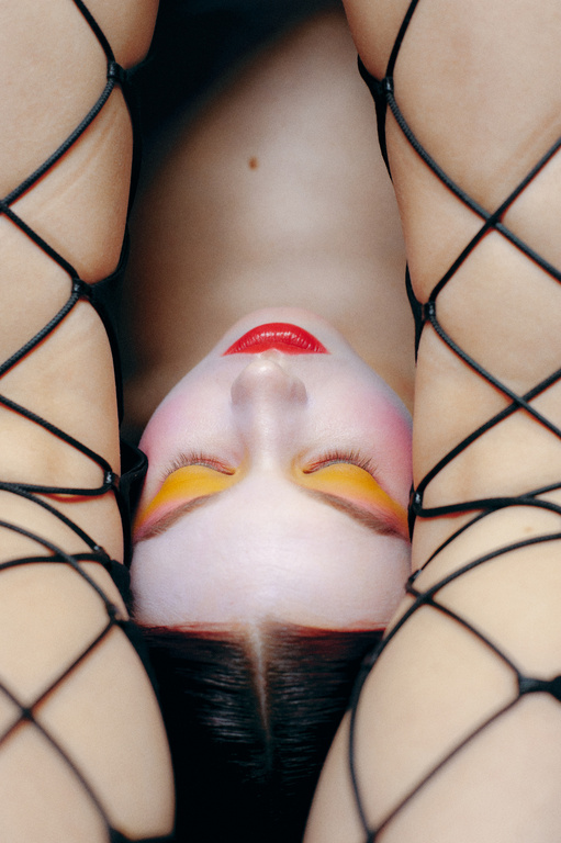 Upside-down view of a fair-skinned person with red lipstick and yellow eyeshadow surrounded by legs wearing black fishnets.