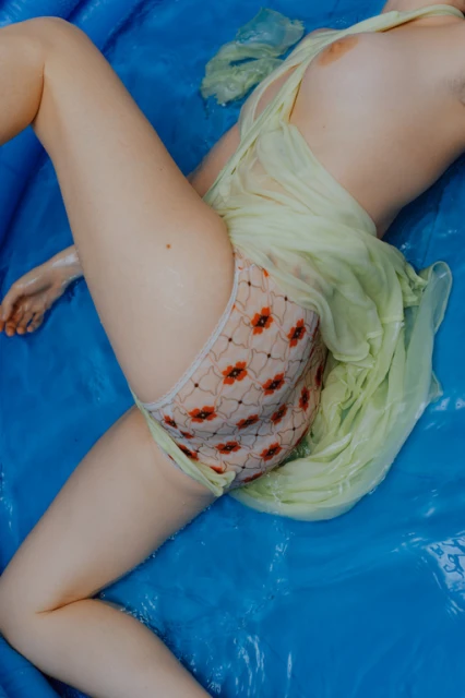 Woman in patterned underwear and a light green sheer dress lies in blue water. Bright overhead studio lighting creates a clean, modern aesthetic.