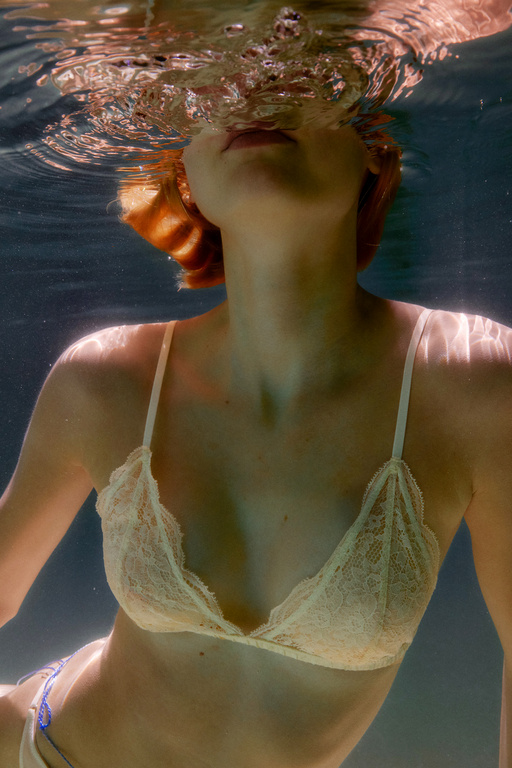 Underwater portrait of a red-haired woman in a white lace bikini top, looking up towards the light through the water's surface.