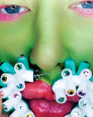 Close-up portrait of a person with green face paint, pink eyeshadow, blue eyes, red lips, and artistic earrings with colorful gems.