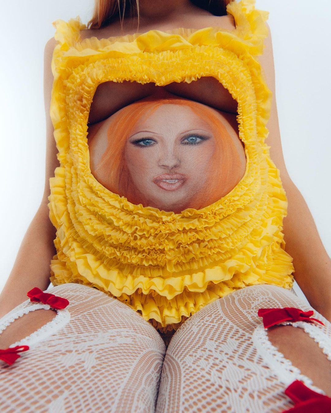 Blonde female model wears a yellow ruffled top with a painted face on her stomach, paired with white fishnet tights and red-bowed heels.