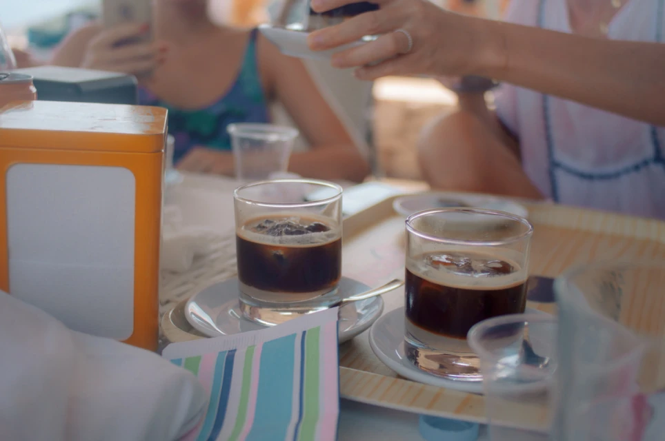 Two glasses of iced coffee sit on a table with other drinks and guests in the background, creating a summery, social atmosphere.