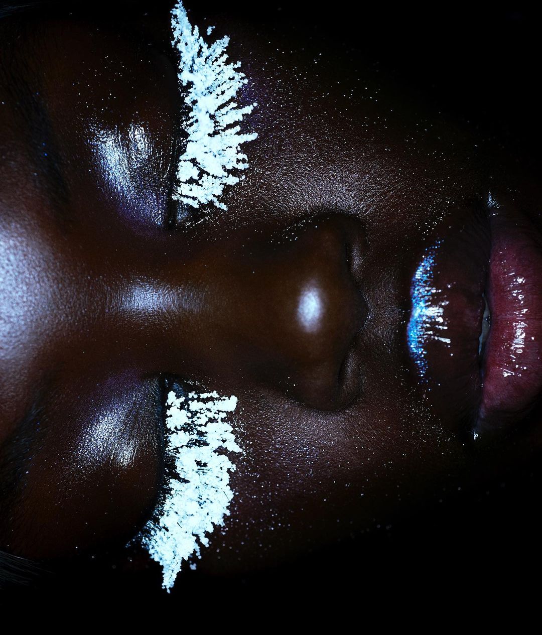 Close-up portrait of a Black woman with makeup, featuring a dark complexion, white cosmetic accents on the eyelids, and reflective lips.
