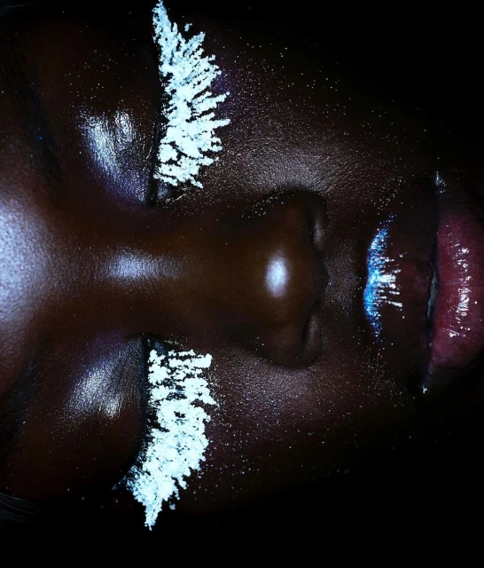 Close-up portrait of a Black woman with makeup, featuring a dark complexion, white cosmetic accents on the eyelids, and reflective lips.