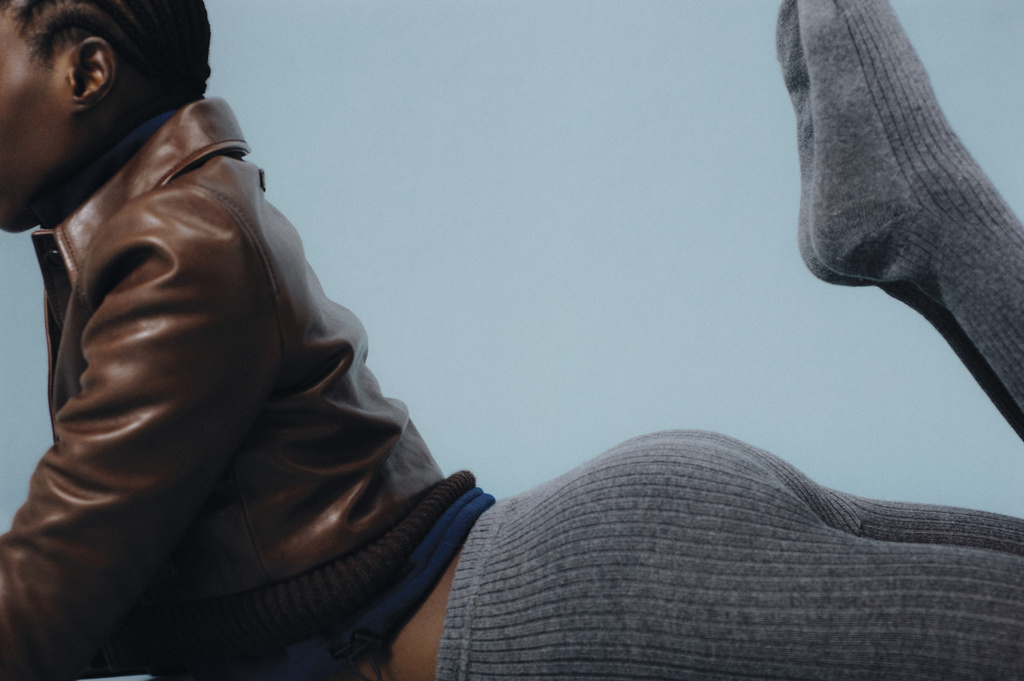 A woman with dark skin and braided hair lies on a light blue surface wearing a brown leather jacket and gray ribbed skirt and socks.