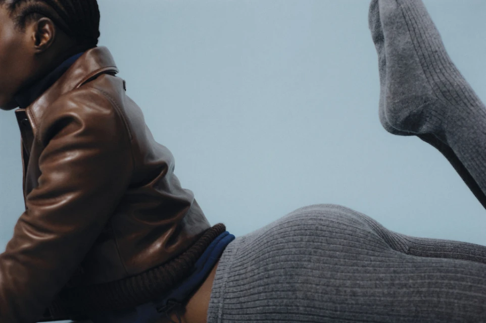 A woman with dark skin and braided hair lies on a light blue surface wearing a brown leather jacket and gray ribbed skirt and socks.