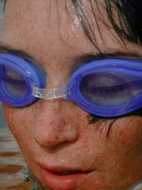 Close-up of a child wearing blue swim goggles. The face is wet with water droplets and has freckles. The background is blurred water.