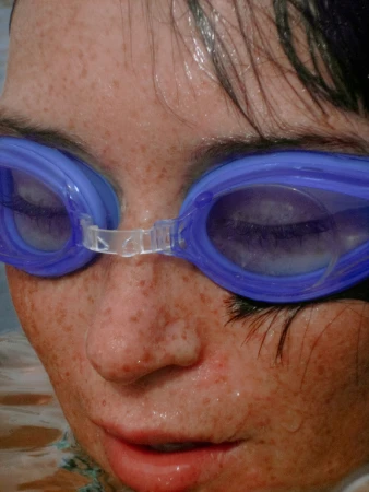 Close-up of a child wearing blue swim goggles. The face is wet with water droplets and has freckles. The background is blurred water.