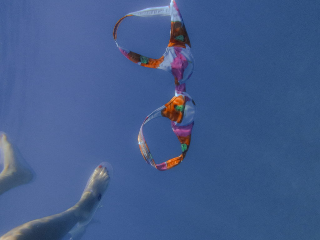Underwater view of a bra floating with a leg and foot of a person swimming by in the background against a blue water backdrop.