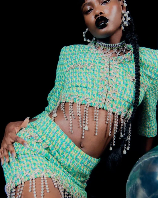 Fashion shot of an adult female wearing a coordinated green tweed skirt and top set with crystal fringe, posing against a black backdrop.