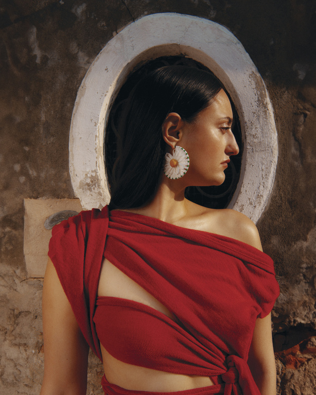 Brunette woman in profile wearing a red dress and daisy earrings, posing near a round window in a textured wall. Fashion shot.