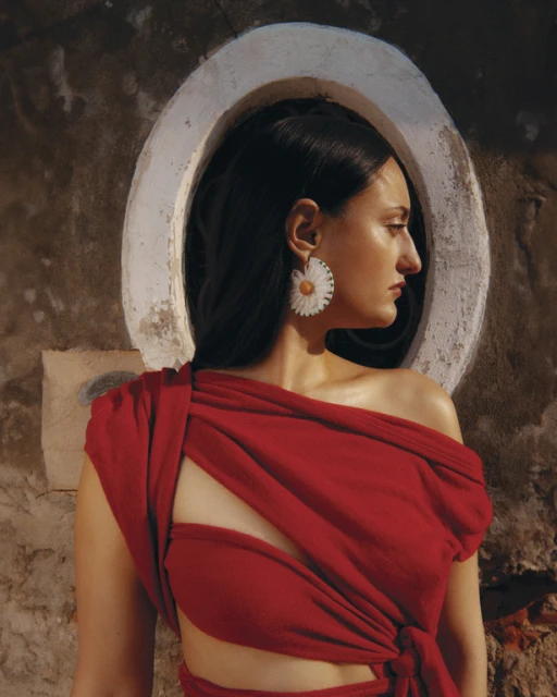 Brunette woman in profile wearing a red dress and daisy earrings, posing near a round window in a textured wall. Fashion shot.