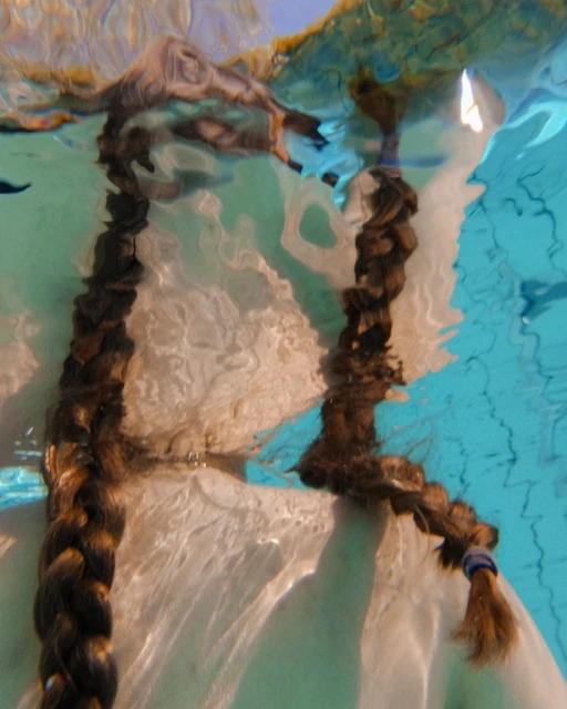 Underwater shot of two dark brown braided pigtails cascading vertically in clear, pale turquoise water with dappled sunlight.
