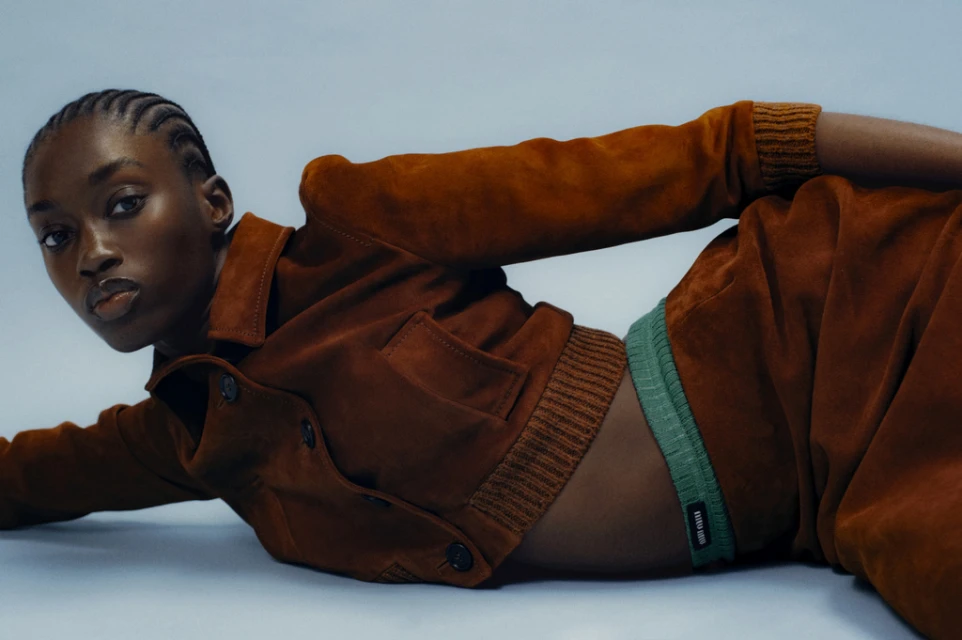 Fashion shot of a Black female model in a brown jacket and wide-leg trousers, lying on a light blue studio backdrop, looking at the camera.