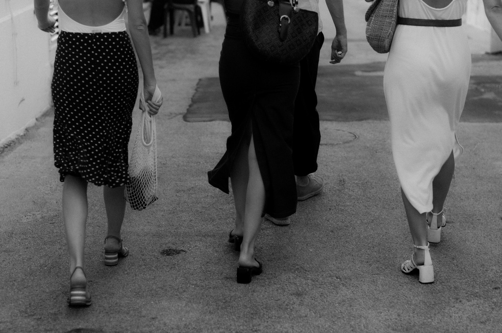 Three women walk together in black-and-white, the one on the left wearing a polka-dot skirt and carrying a mesh bag.