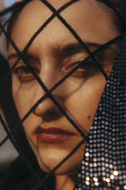 Close-up portrait of a woman with a black net veil. The focus is on her face, with a dark sequin fabric visible on the side.