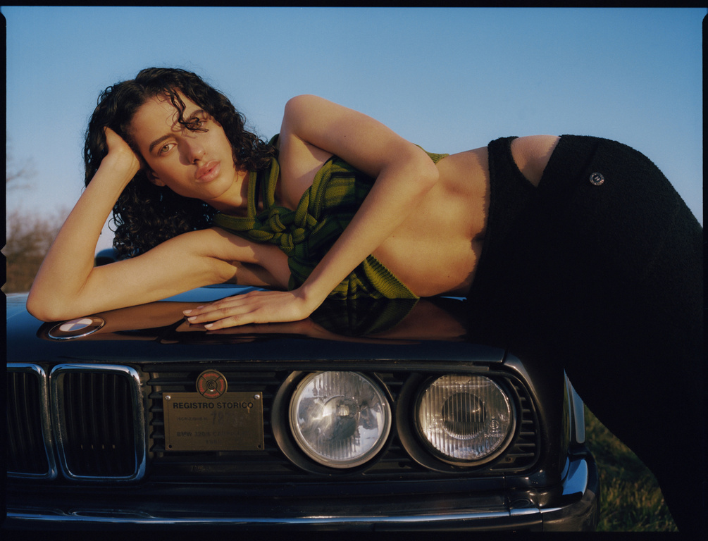 A woman with curly dark hair and green and yellow scarf lies on the hood of a black car under a bright blue sky, with greenery in the background.