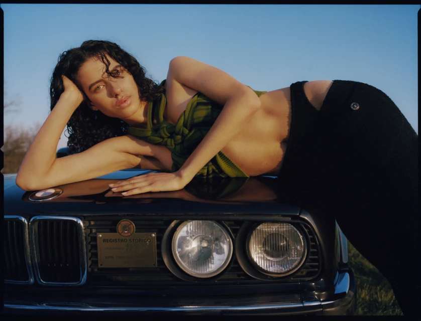A woman with curly dark hair and green and yellow scarf lies on the hood of a black car under a bright blue sky, with greenery in the background.